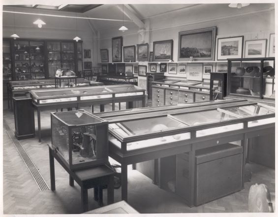 A black and white photograph of littlehampton museum with wooden cases. There is archaeology and natural history on display.